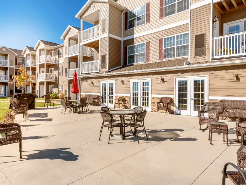 Outdoor patio area of a senior living facility with multiple round tables and chairs, wicker seating, a red umbrella, and a barbecue grill. The building has beige siding with white trim and multiple balconies and windows.