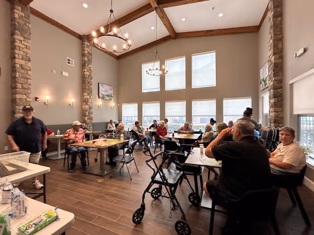 A spacious dining area with seniors at tables, walkers nearby, and warm pendant lighting.