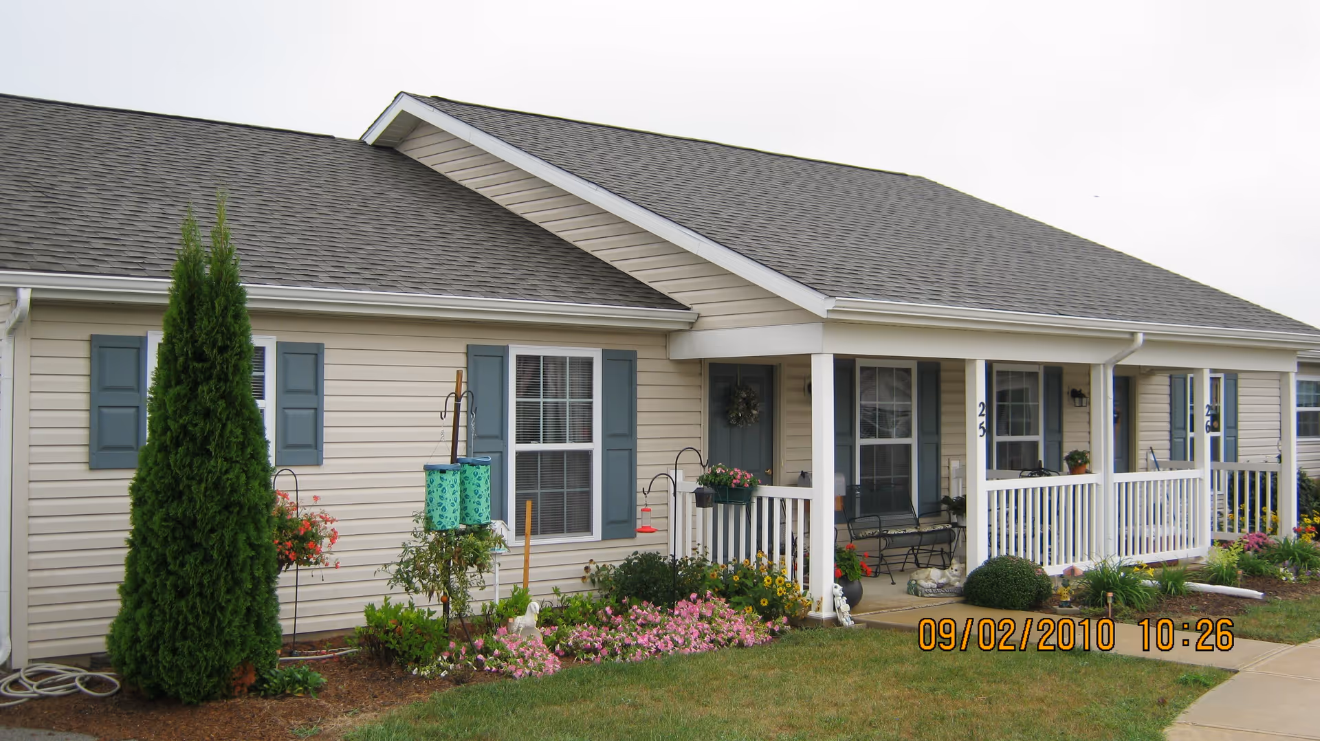 Single-story light-colored building with a covered front porch, white railing, and landscaped flowerbeds.