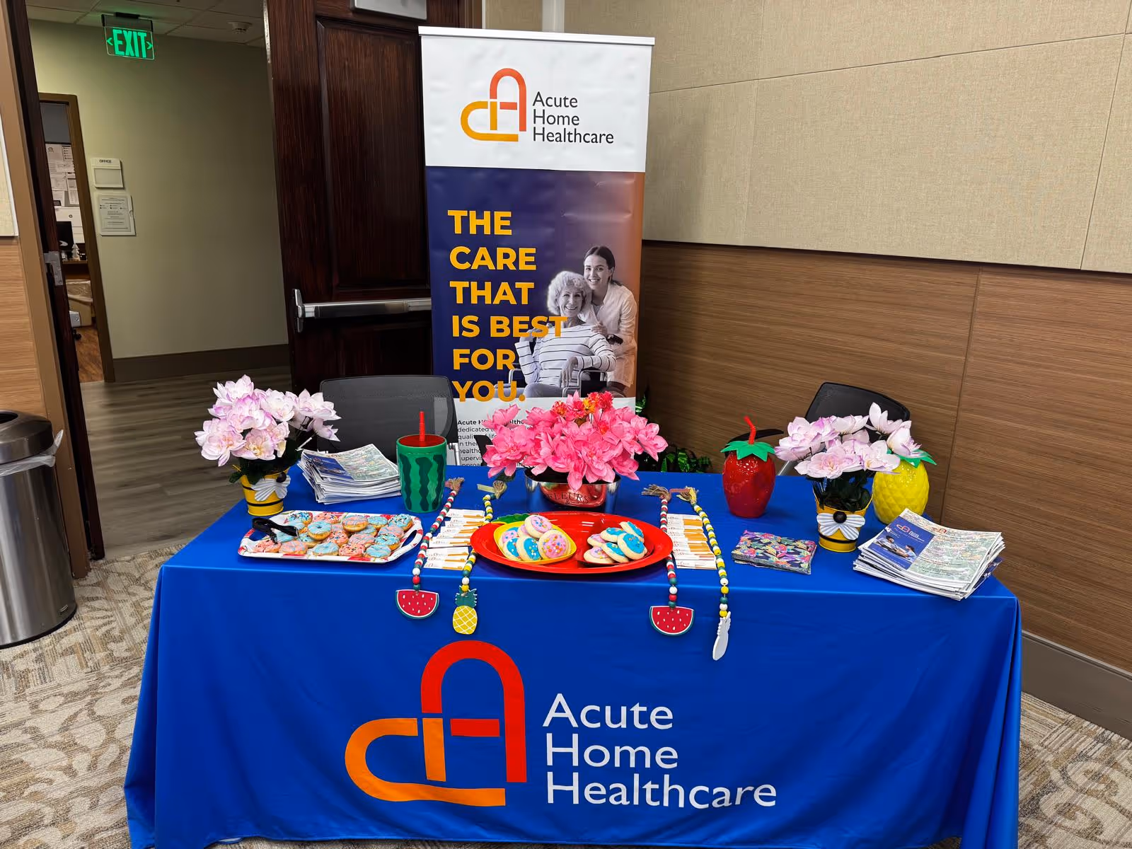 Promotional table with blue cloth and flowers inside a hallway, Acute Home Healthcare banner behind