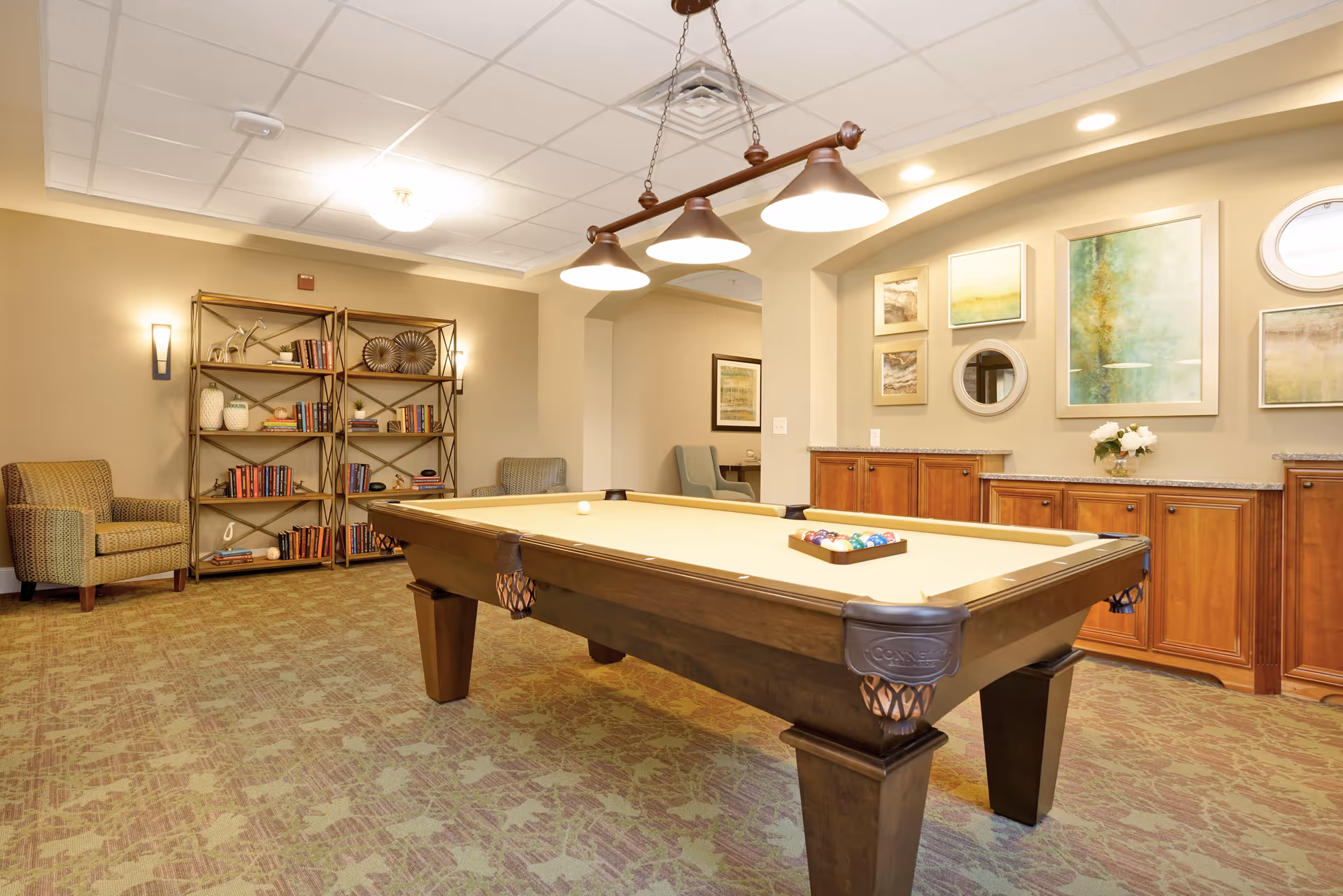 A well-lit recreational room featuring a pool table with balls arranged for a game. The room has beige walls adorned with framed artwork and a round mirror. There are wooden cabinets along one wall, a bookshelf filled with books and decorative items, and comfortable armchairs placed around the room. Overhead, a light fixture with three lamps hangs above the pool table.