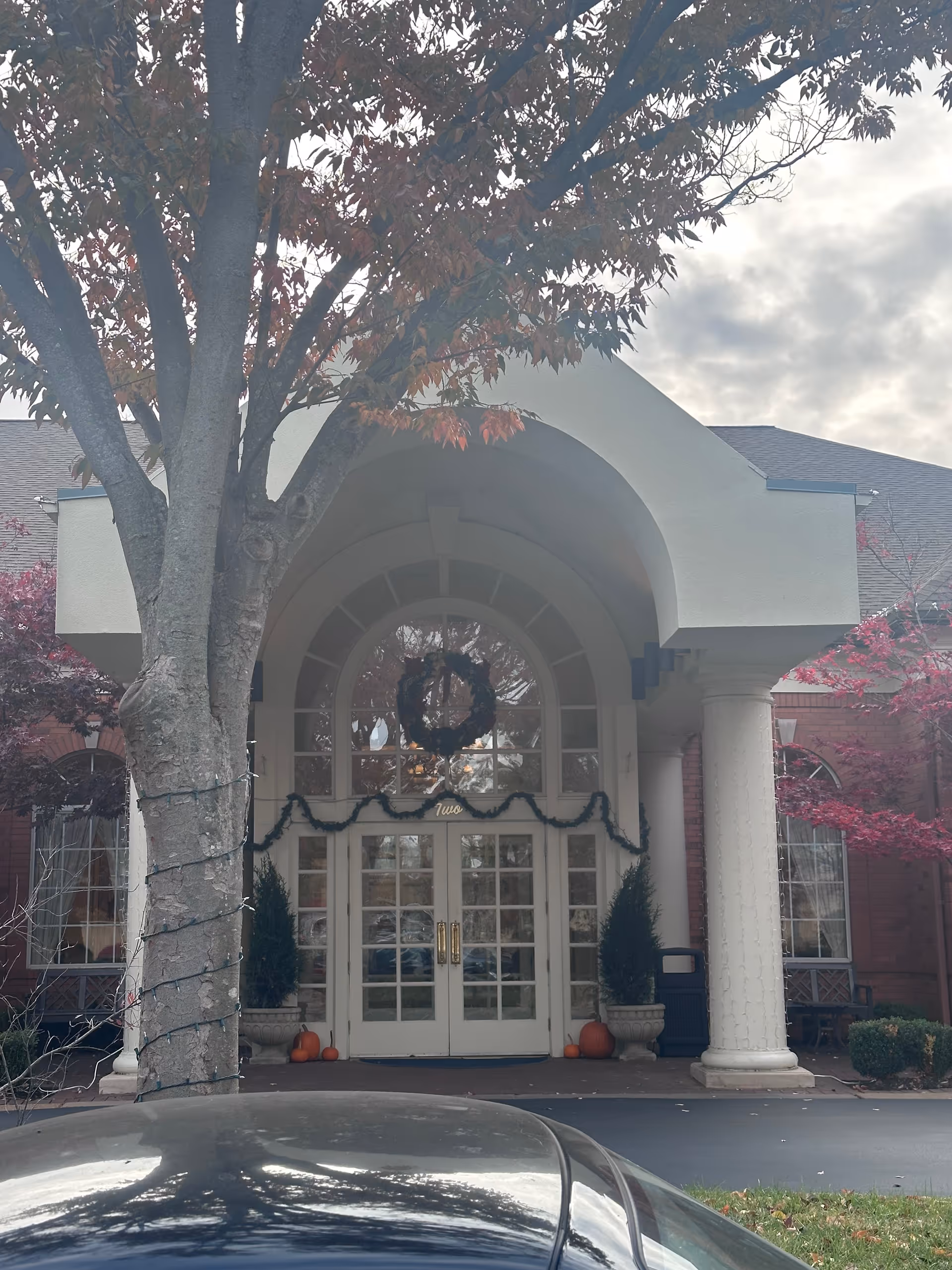 Front entrance of a senior care building with an arched portico, double glass doors, a wreath, flanking columns, and a tree in the foreground.