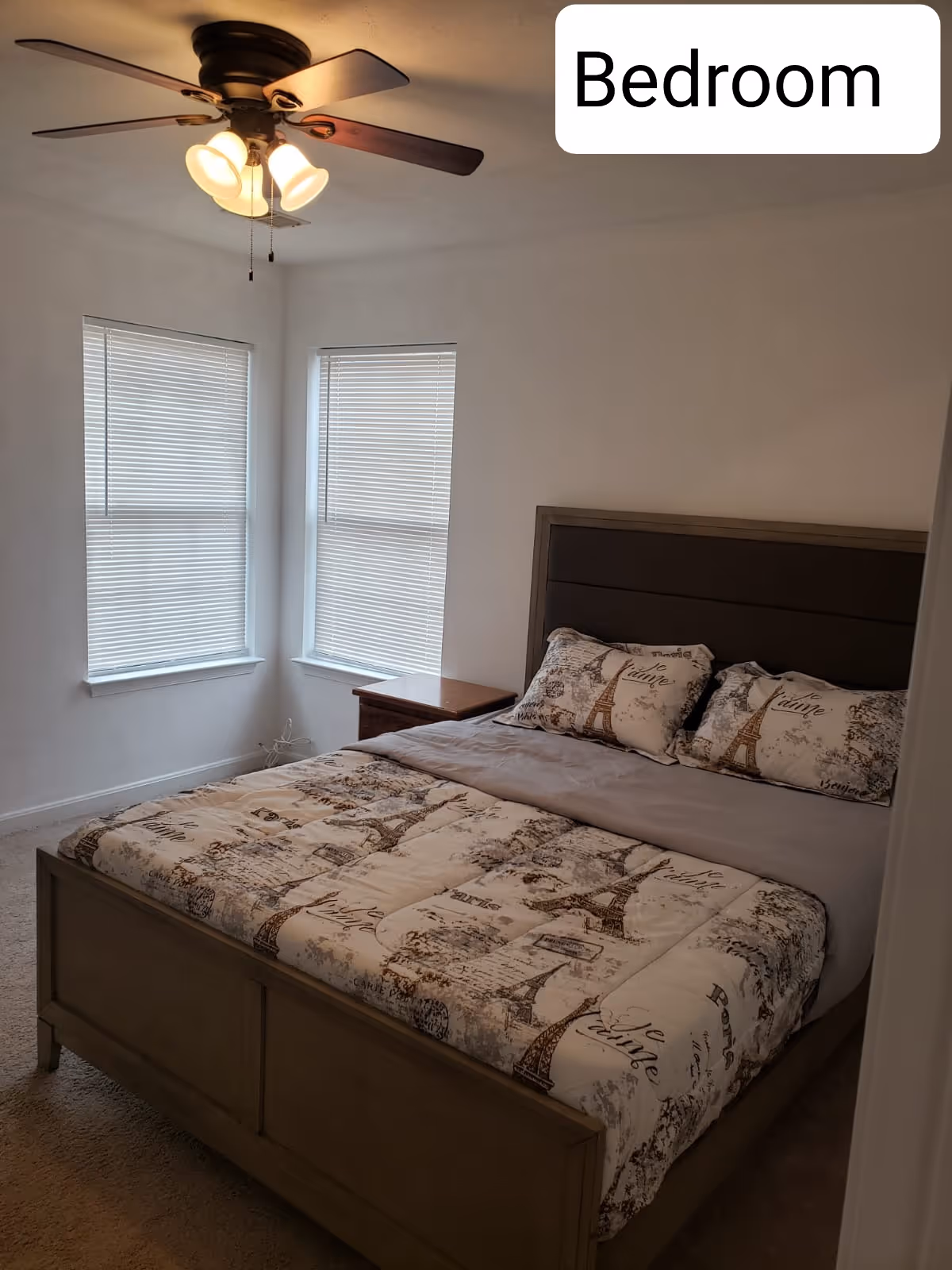 A senior living bedroom with a wooden bed, two windows with blinds, and a ceiling fan.