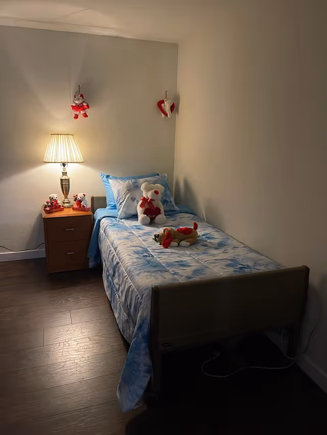 Cozy single bedroom with blue bedding, teddy bears, and a nightstand beside a lamp.