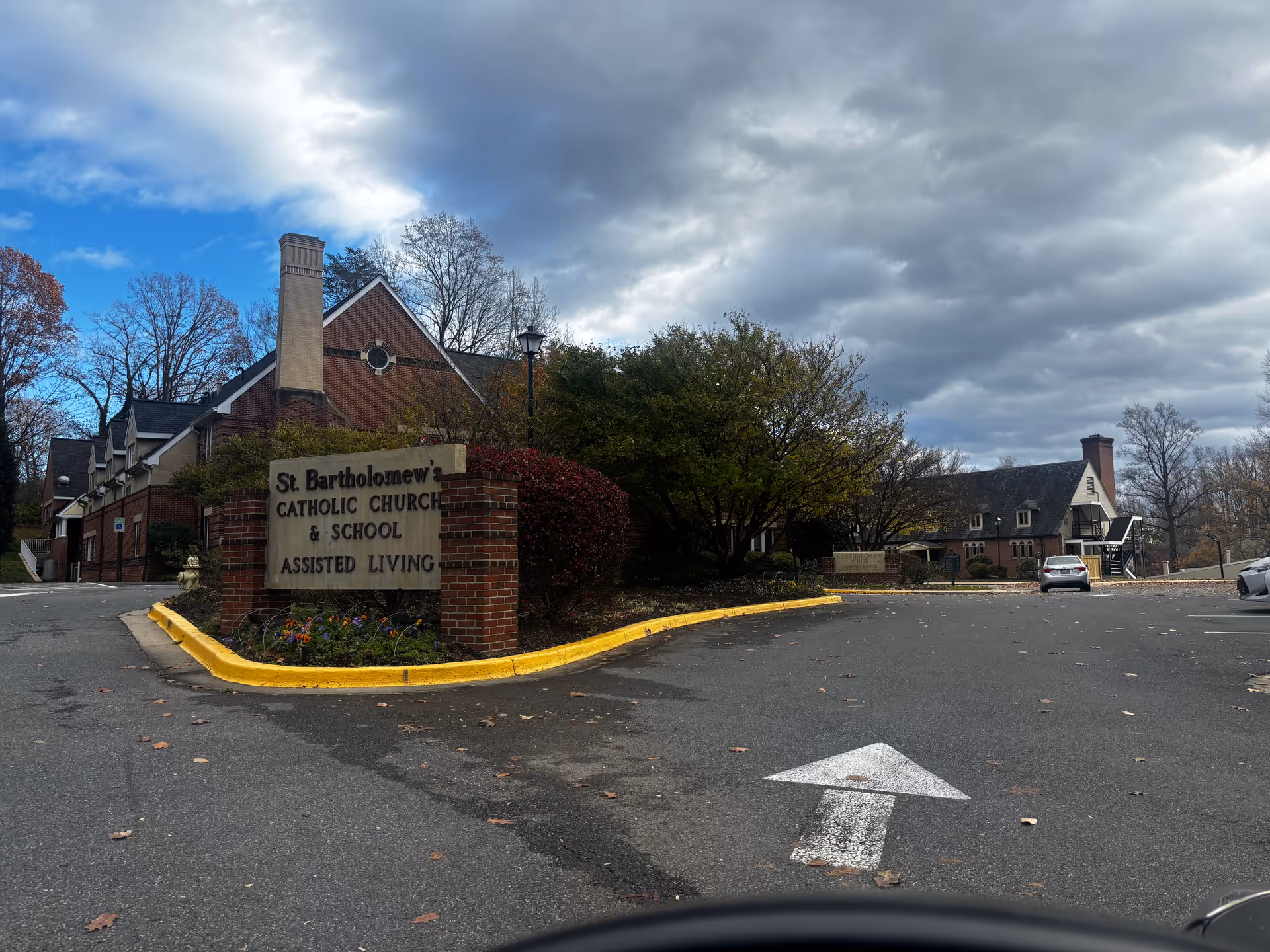 Brick entrance sign reading 'St. Bartholomew's Catholic Church & School Assisted Living' with brick buildings and a cloudy sky behind it.