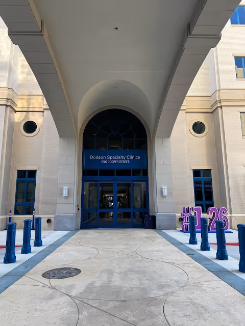 Front entrance of a beige clinic with an arched doorway and blue-framed windows.