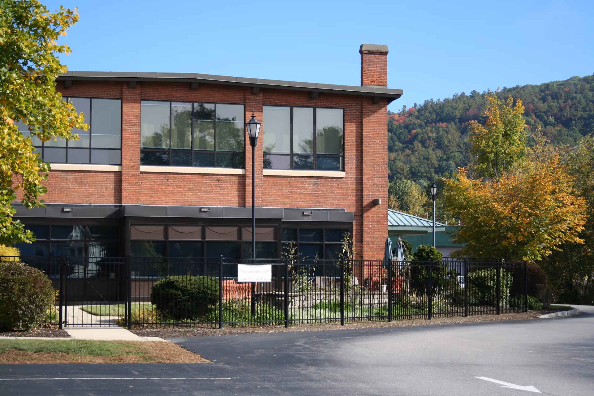 Brick senior care facility building with large windows, black fence, and autumn trees.