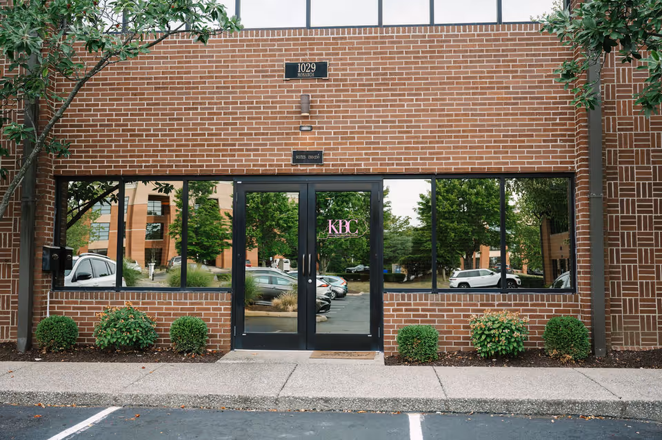 Brick building facade with glass doors, greenery, and reflections in windows.