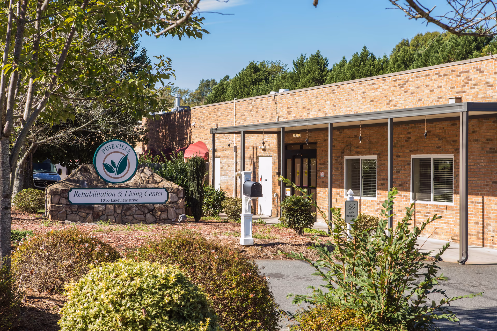 Front entrance of Pineville Rehabilitation & Living Center showing a stone sign, mailbox, and landscaped shrubbery.