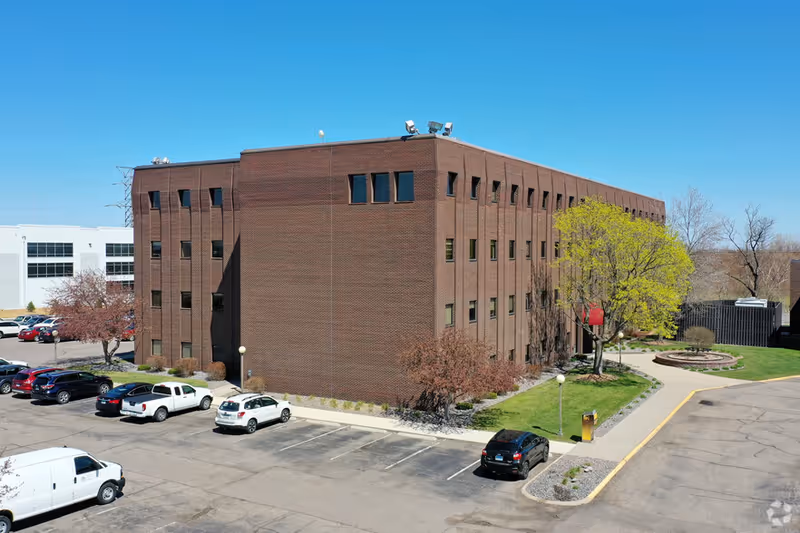 A brick multi-story facility with a parking lot and landscaped courtyard under a clear blue sky.