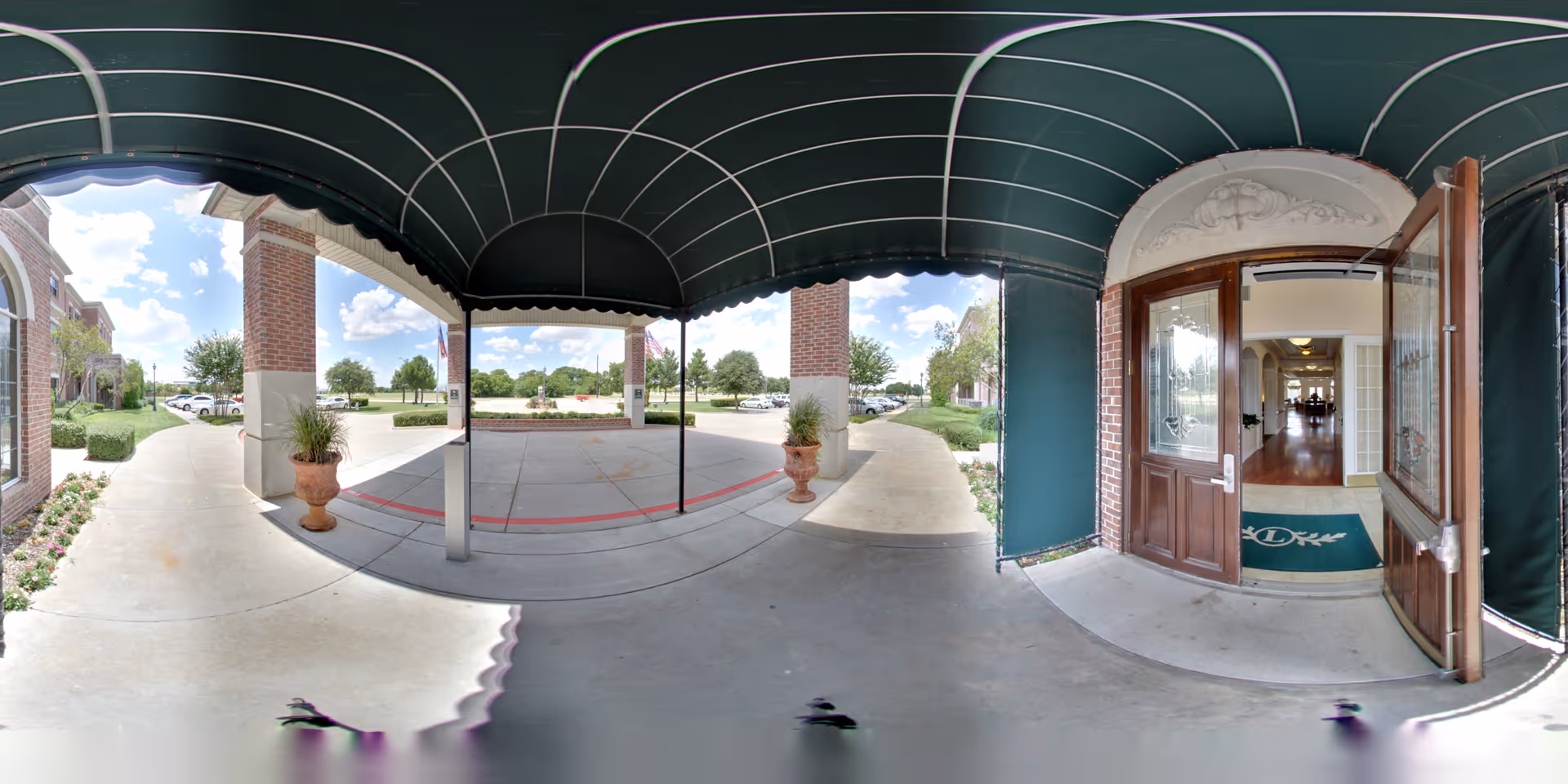 Covered entrance area of a senior living facility with a green canopy overhead, brick columns, potted plants, and a wooden door open to an interior hallway with wooden floors and a green mat with a logo.