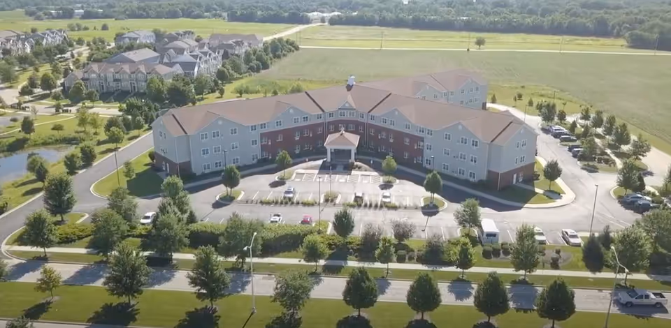 Aerial view of Heritage Woods of Plainfield, a large senior living facility with a three-story building surrounded by parking lots, trees, and green fields.