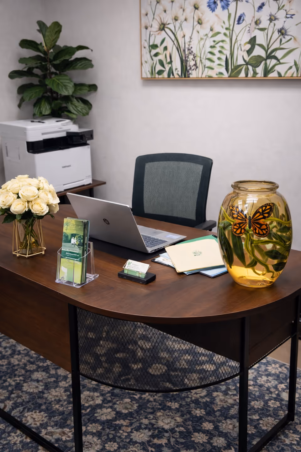 Office desk with laptop, flowers, butterfly jar, brochures, and printer.