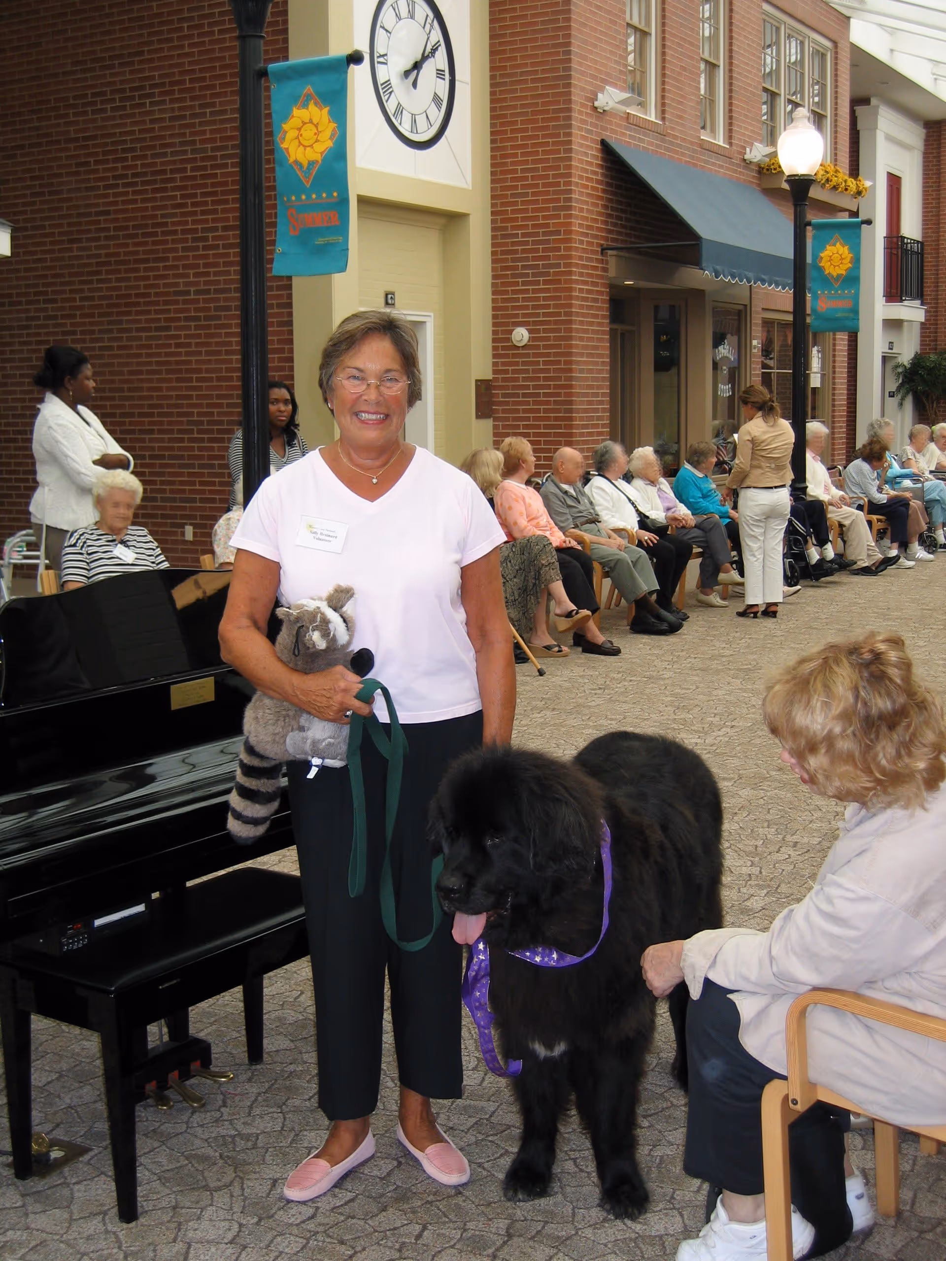 A smiling woman holding a stuffed raccoon toy and a leash attached to a large black dog with a purple collar stands indoors in front of a black piano. Several elderly people are seated along the wall in the background, and another woman is interacting with the dog. The setting appears to be a communal area with brick walls, a clock, and banners.