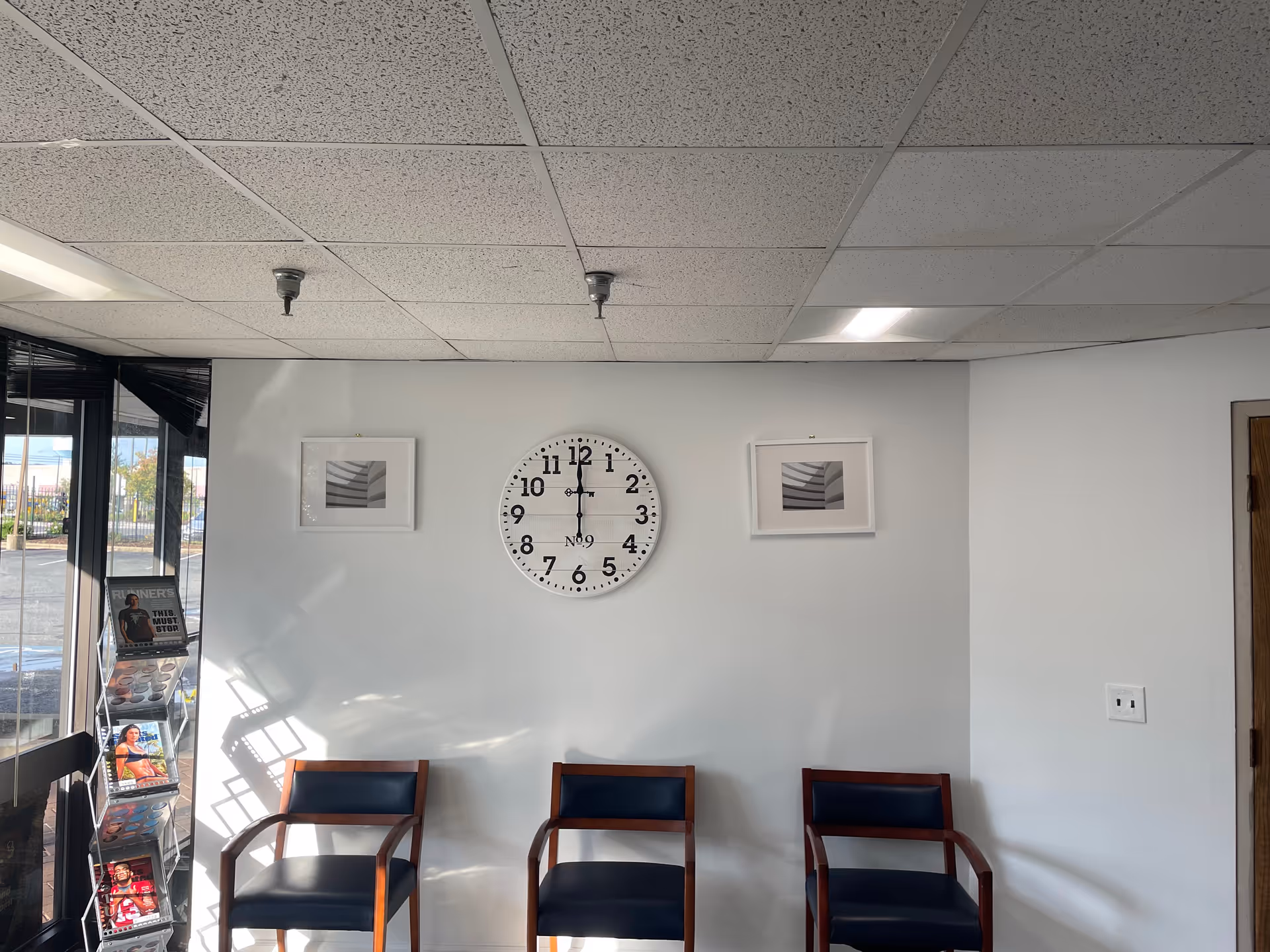 Waiting area inside a facility with four wooden and blue chairs, a wall clock, and magazines.