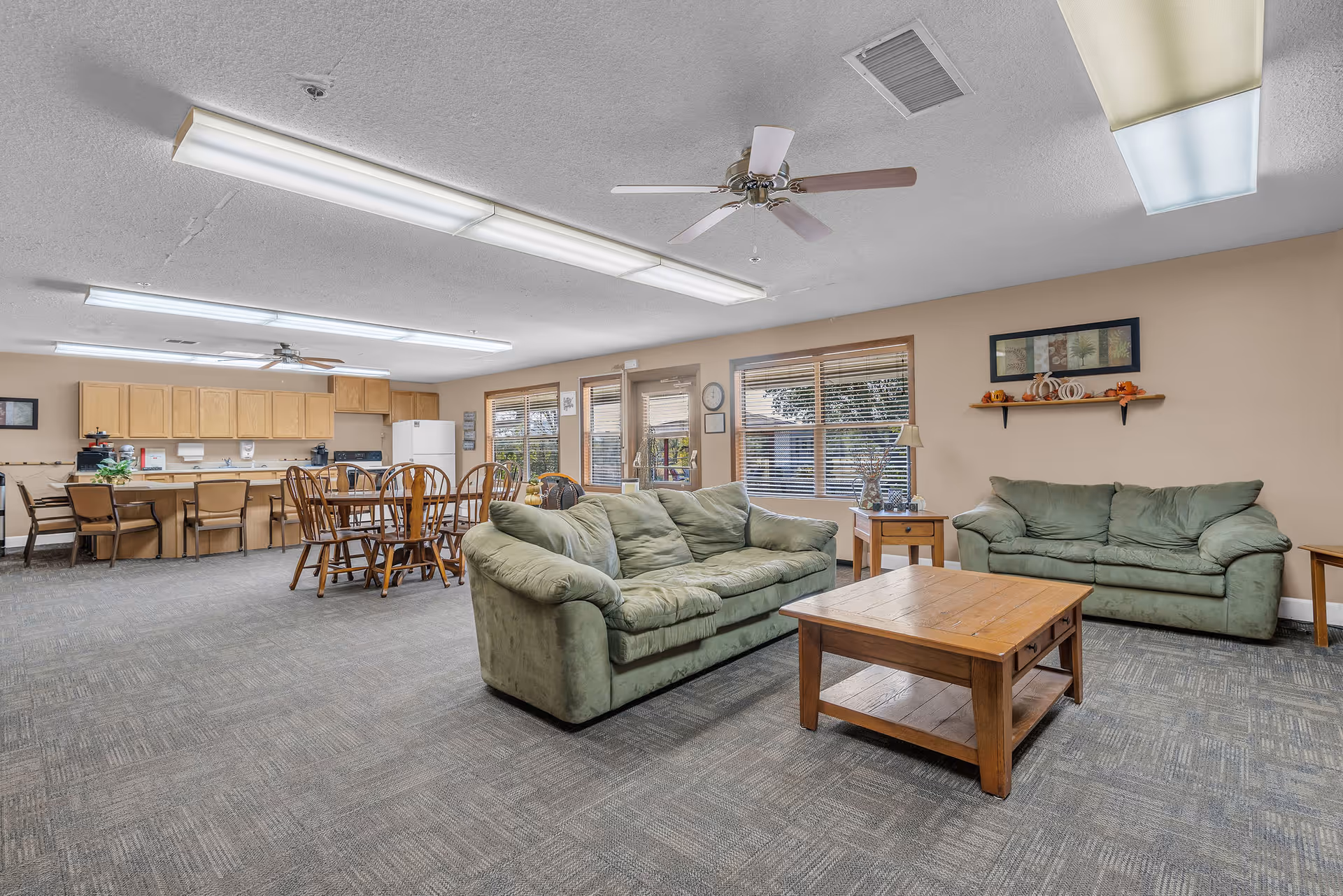 Spacious senior living lounge with green sofas, coffee table, and adjacent dining/kitchen area.
