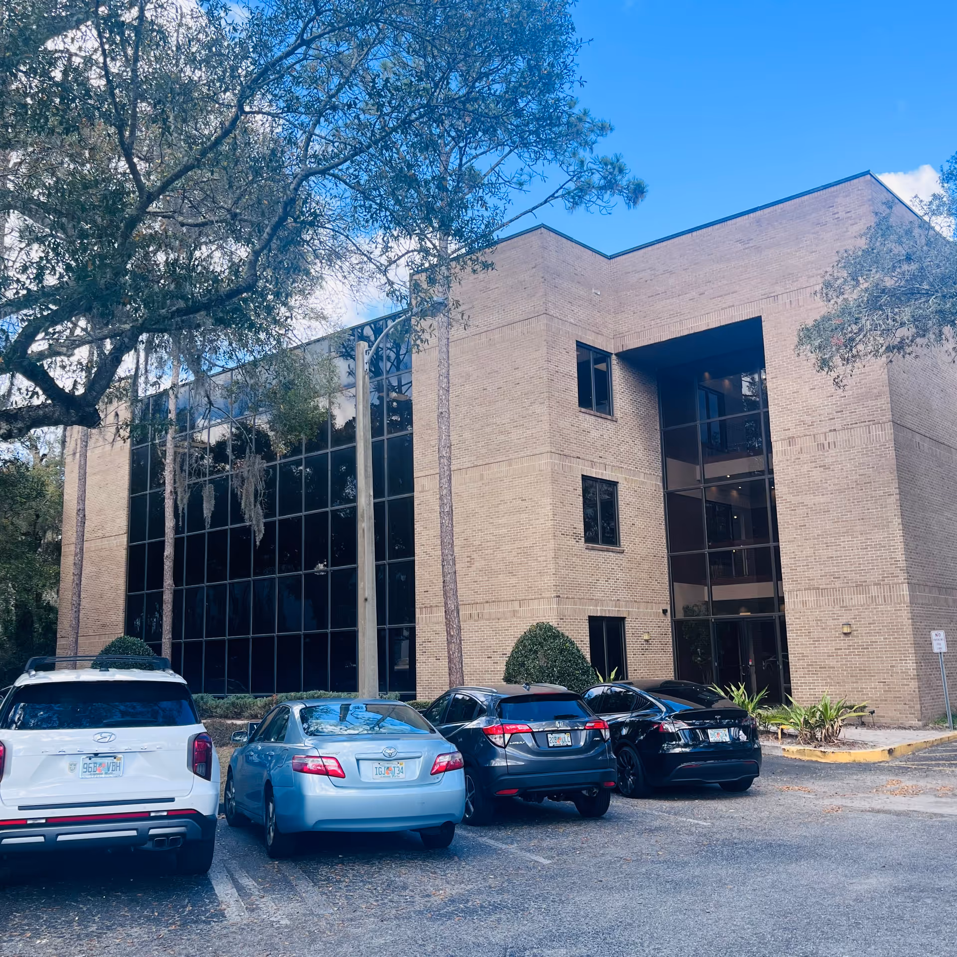 Brick senior living facility exterior with large glass windows, trees, and a parking lot.