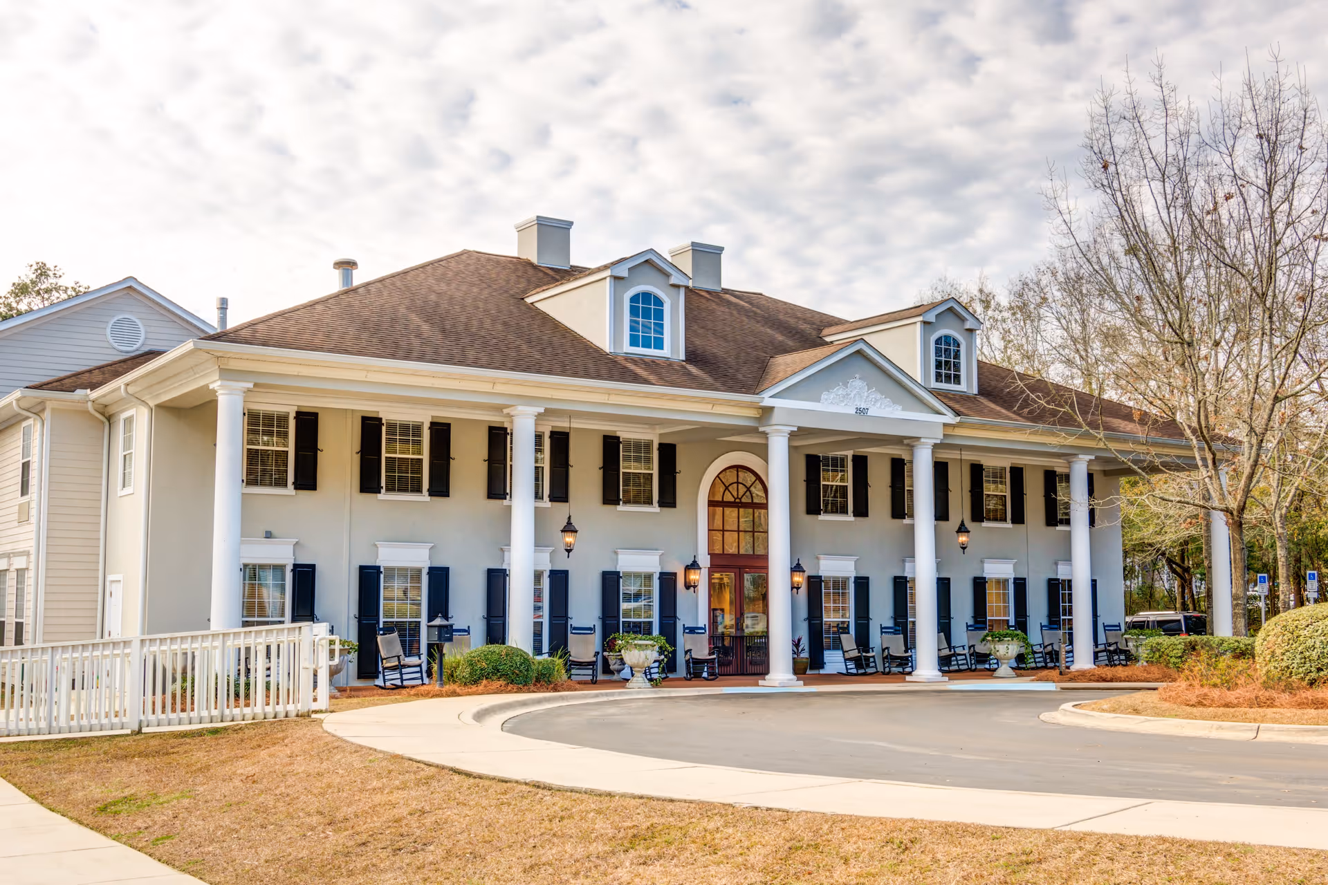 Front exterior view of a large two-story building with white columns, black shutters, and a brown roof. The entrance has a rounded arch door with glass panels and is flanked by outdoor lanterns. There are several rocking chairs on the porch and a circular driveway in front. Trees and bushes surround the building under a partly cloudy sky.