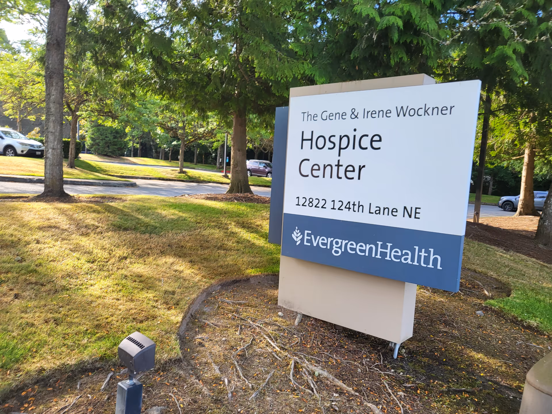 Outdoor sign reads The Gene and Irene Wockner Hospice Center at EvergreenHealth, near trees and cars.