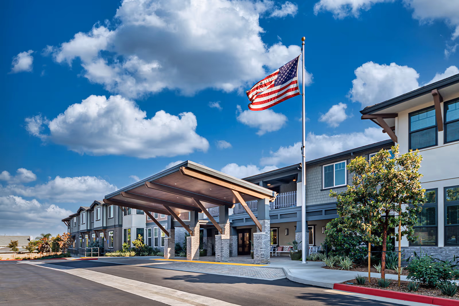 Exterior view of Allara Senior Living facility showing a large covered entrance with stone pillars, an American flag on a flagpole, and a two-story building under a blue sky with scattered clouds.