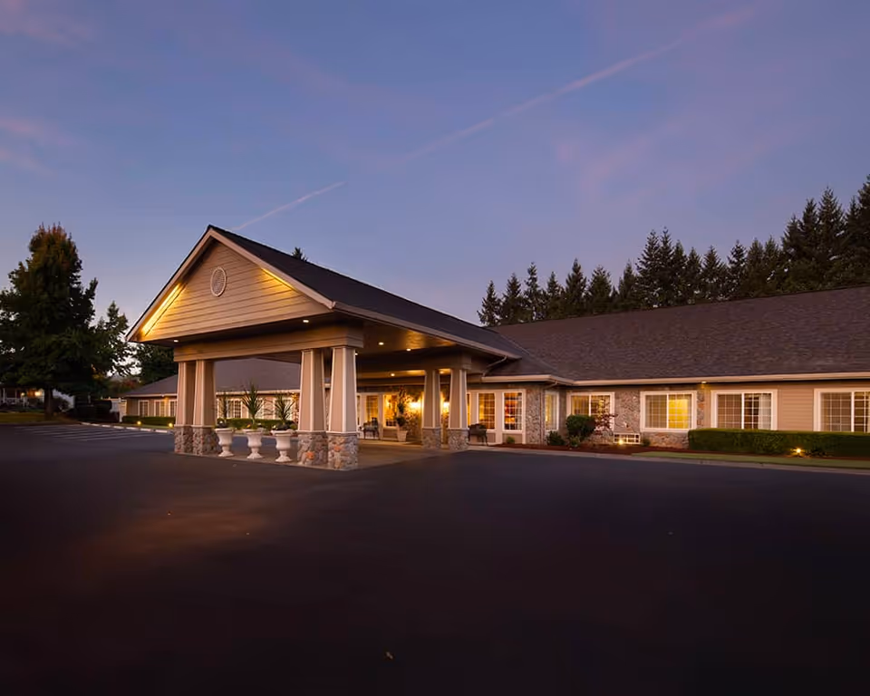 Dusk view of a single-story senior living facility's front entrance with a covered porte-cochère and illuminated windows.