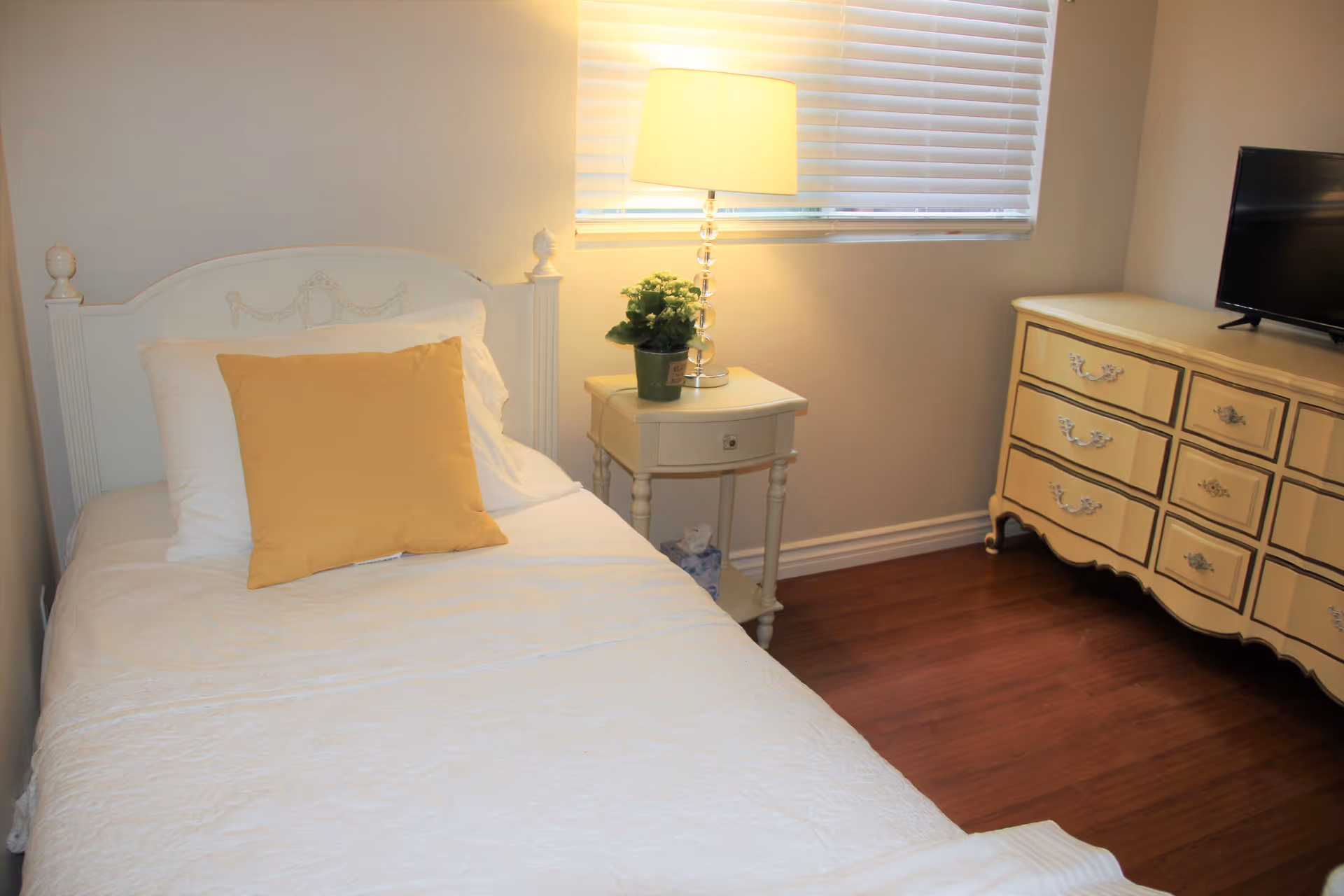 A cozy bedroom with a single white bed featuring a yellow pillow and white bedding. Next to the bed is a small white nightstand with a potted plant and a lamp with a white shade. A window with white blinds is above the nightstand. Across from the bed is a cream-colored dresser with multiple drawers and a flat-screen TV on top. The room has wooden flooring and light-colored walls.