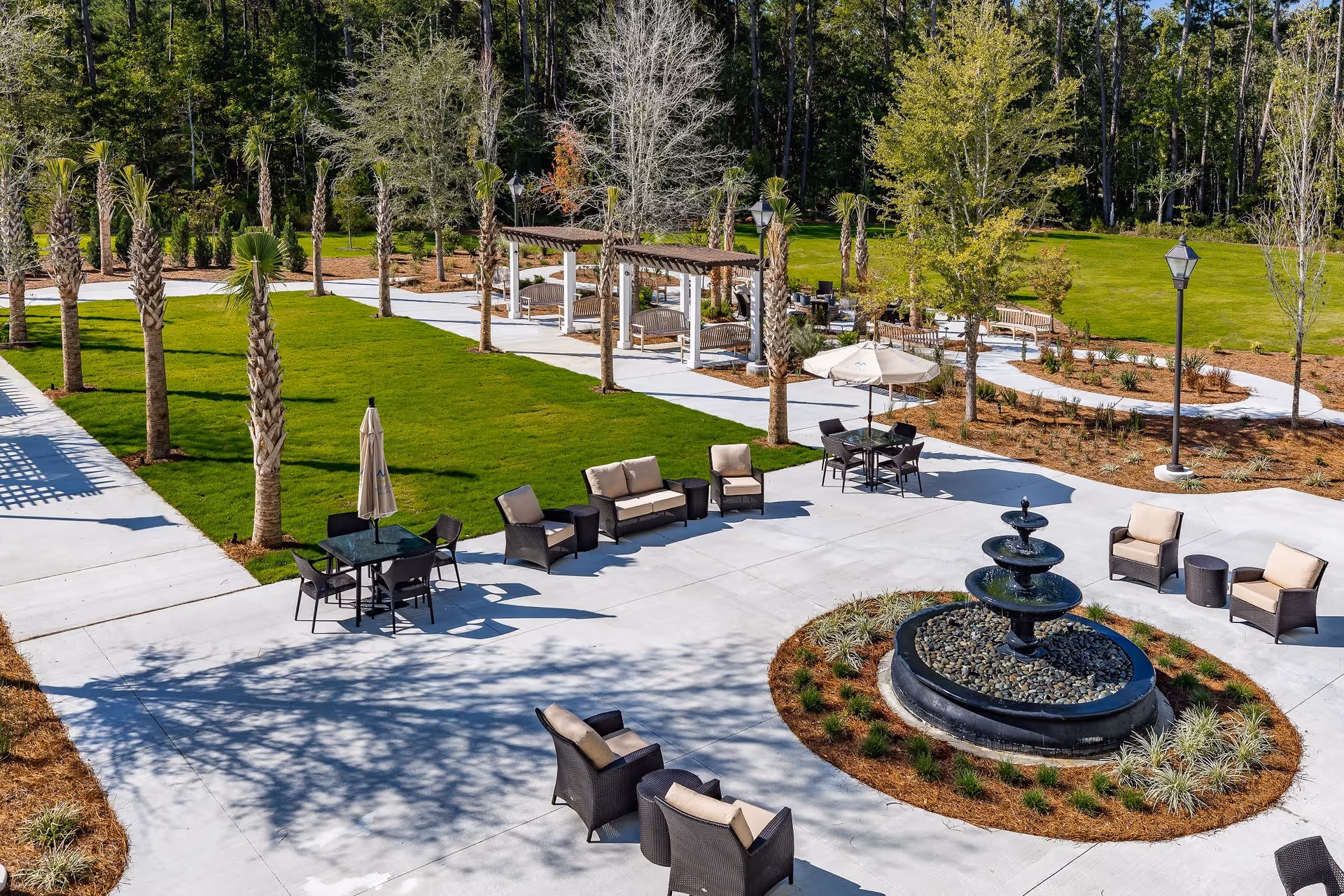 Outdoor patio area with multiple seating arrangements including chairs, sofas, and tables with umbrellas surrounding a tiered water fountain. The area is paved with concrete and bordered by green grass, palm trees, and other landscaping. There are pergolas with benches in the background and a wooded area beyond the lawn.
