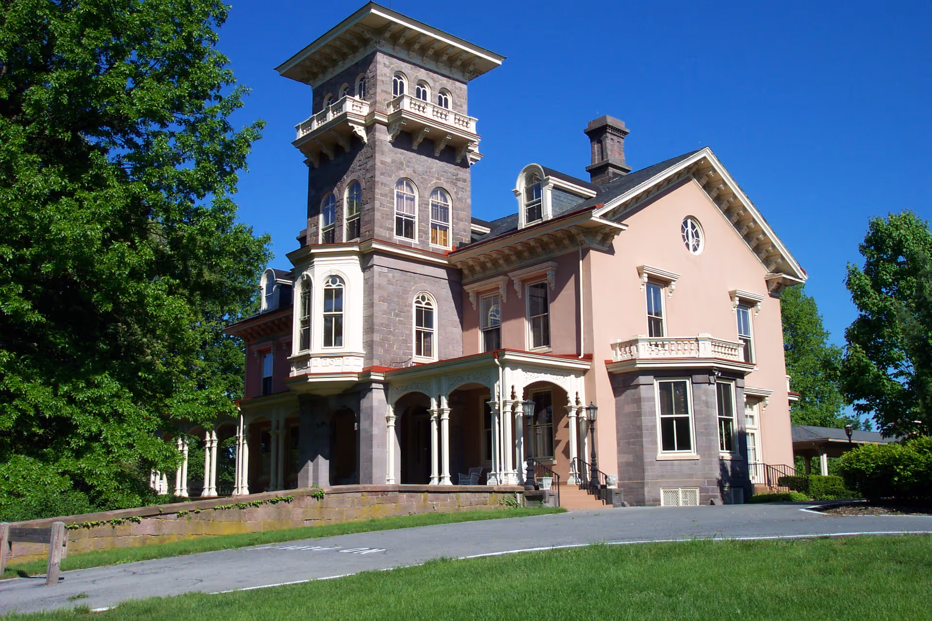 A large Victorian-style manor with a tower, wraparound porch, and columns set on landscaped grounds under a clear blue sky.