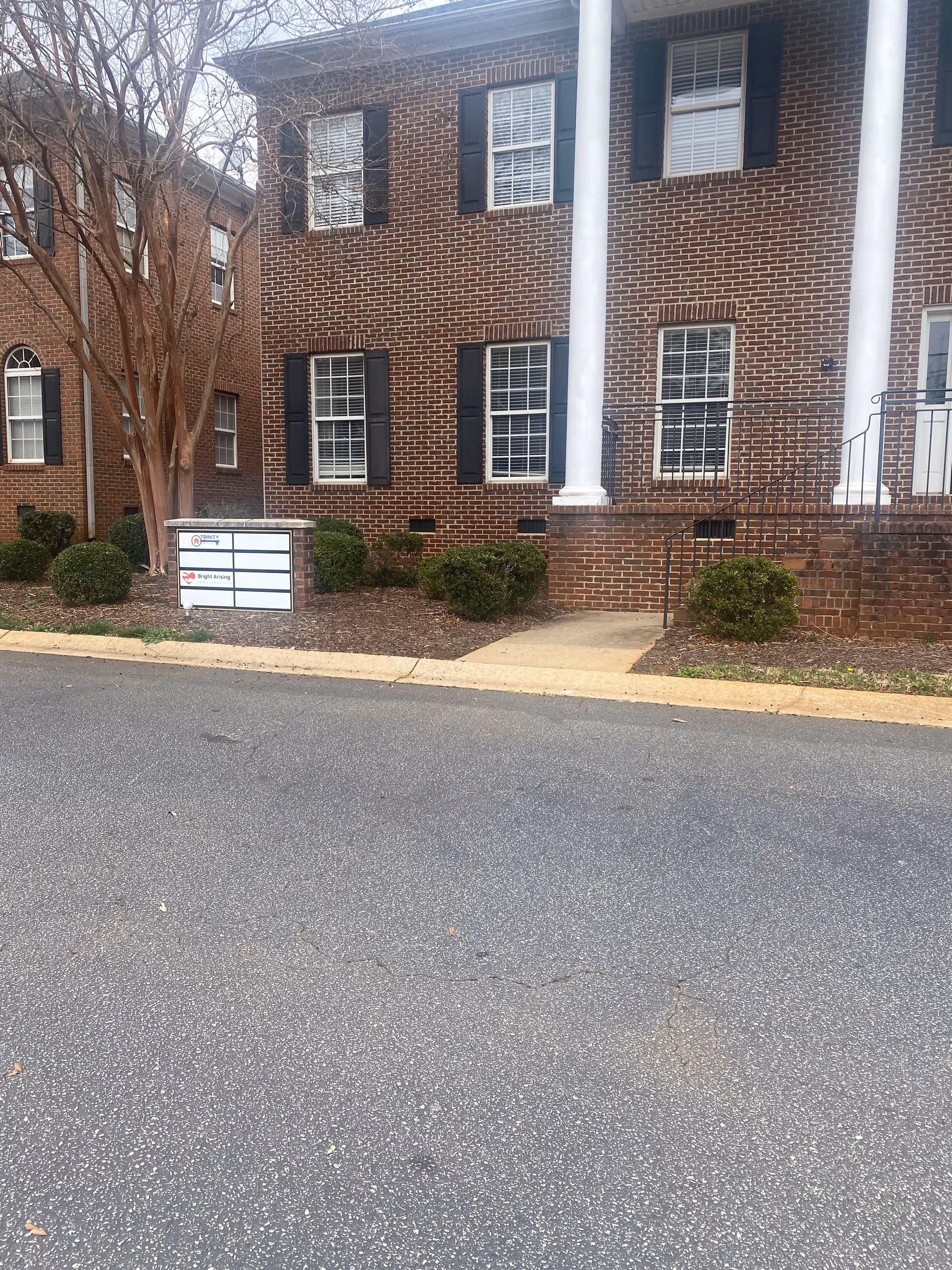 Brick senior living building exterior with white columns, windows, shrubs, and a sign by the curb.