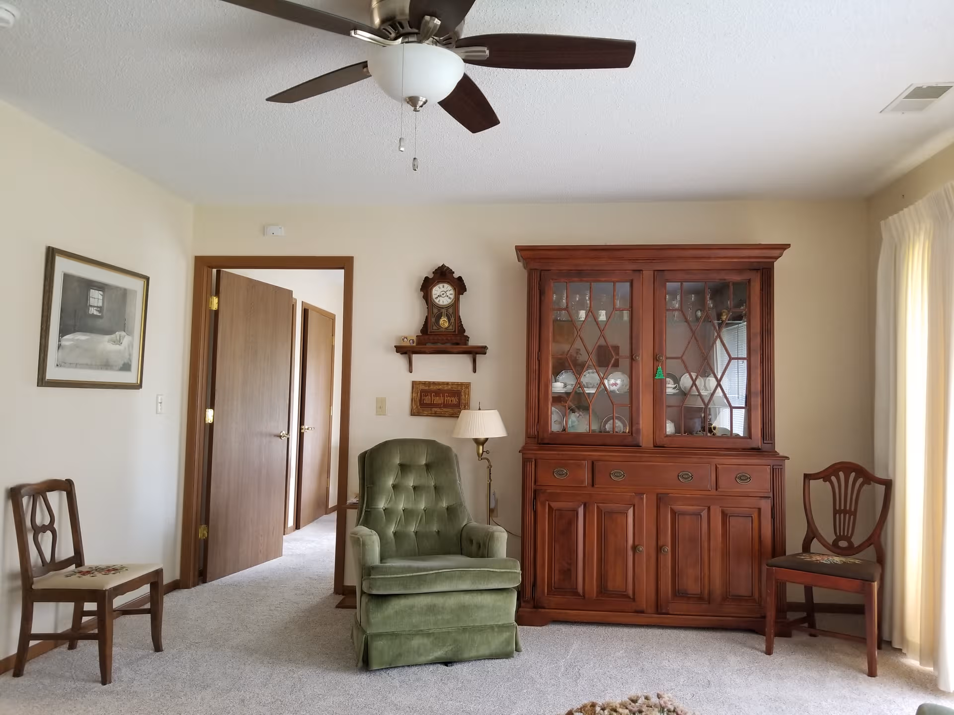 A cozy living room with a green upholstered chair, china cabinet, and ceiling fan.