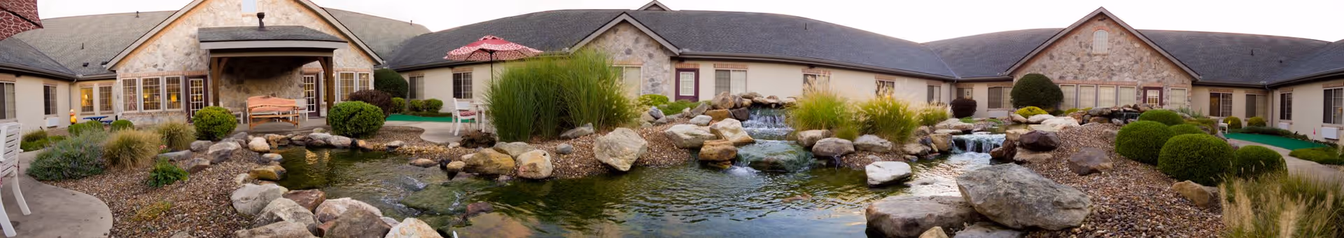 Panoramic exterior courtyard showing a pond with rock waterfalls and landscaping in front of a single-story senior living building.