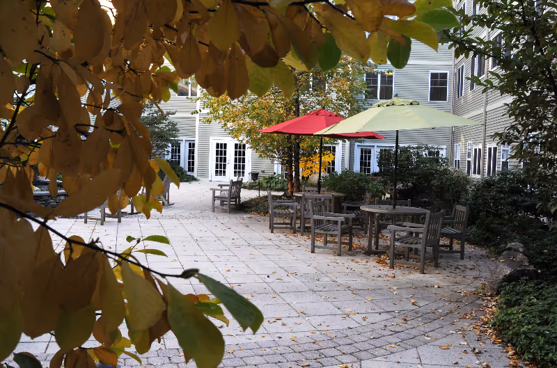 Courtyard patio with outdoor tables and colorful umbrellas beside a multi-story senior living building framed by autumn leaves.