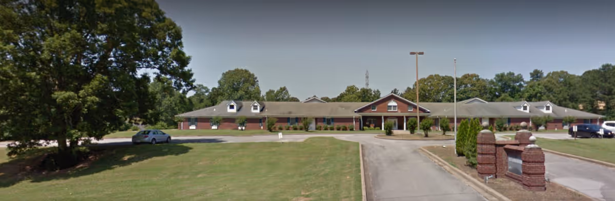 Single-story brick senior living facility with a driveway, lawn, and mature trees.