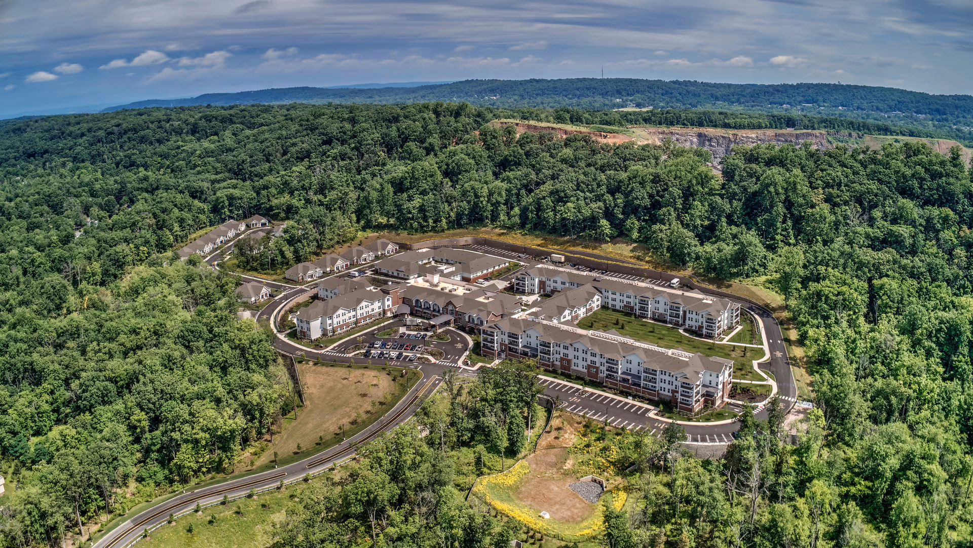 Aerial view of The Delaney of Bridgewater senior living facility surrounded by dense green forest and hills under a partly cloudy sky. The facility consists of multiple connected buildings with parking lots and roads encircling the property.