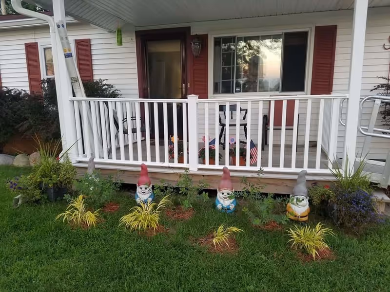 Front porch with a white railing, potted plants and three garden gnomes displayed on the lawn in front of a house.