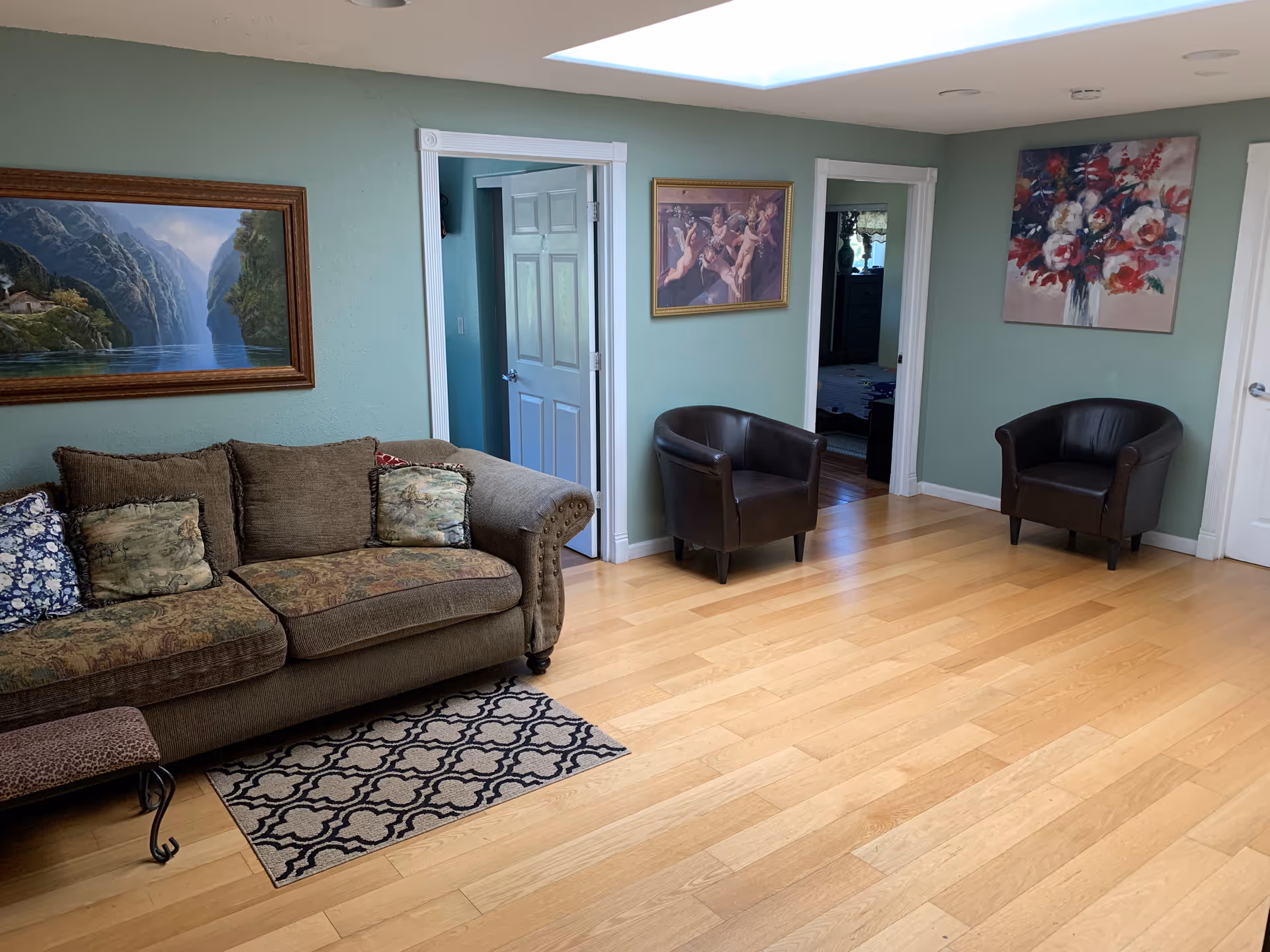 A cozy living room with light green walls and wooden flooring. There is a brown upholstered sofa with patterned cushions on the left, a small patterned rug in front of it, and two dark brown leather armchairs positioned against the walls. Three paintings hang on the walls: a landscape with mountains and water, a classical painting of cherubs, and a colorful floral artwork. Two doorways lead to other rooms.