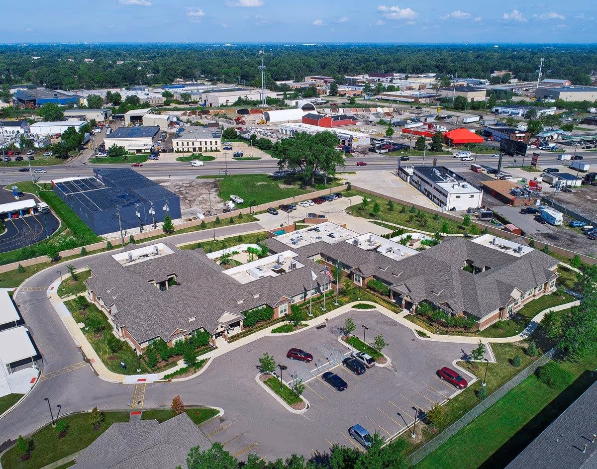 Aerial view of American House Freedom Place Roseville facility showing a large building with multiple wings, surrounded by parking lots and greenery, with a commercial area and roads in the background under a clear blue sky.