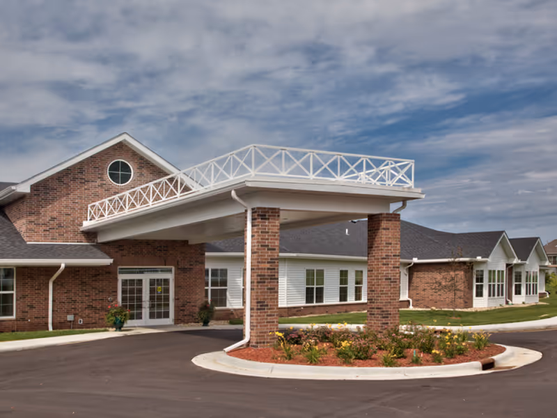 Exterior front entrance of a brick senior living facility featuring a covered drive-through porte-cochère and landscaped circular planter.
