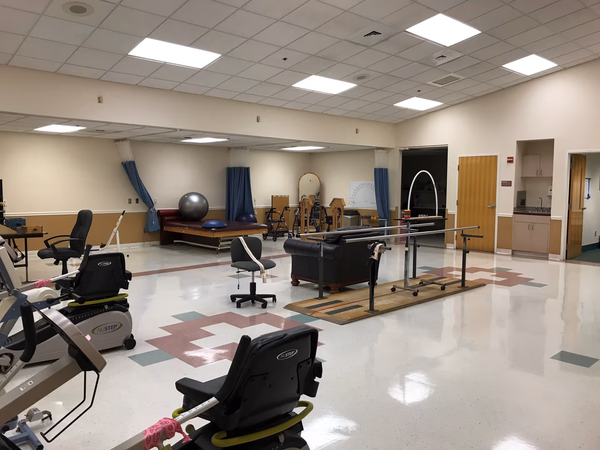 A spacious rehabilitation room with exercise equipment including stationary bikes, parallel bars for walking practice, a therapy table with exercise balls, chairs, and walkers. The room has a tiled floor with a geometric pattern, beige walls, and a drop ceiling with fluorescent lighting. There are several doors and a small kitchenette area visible in the background.