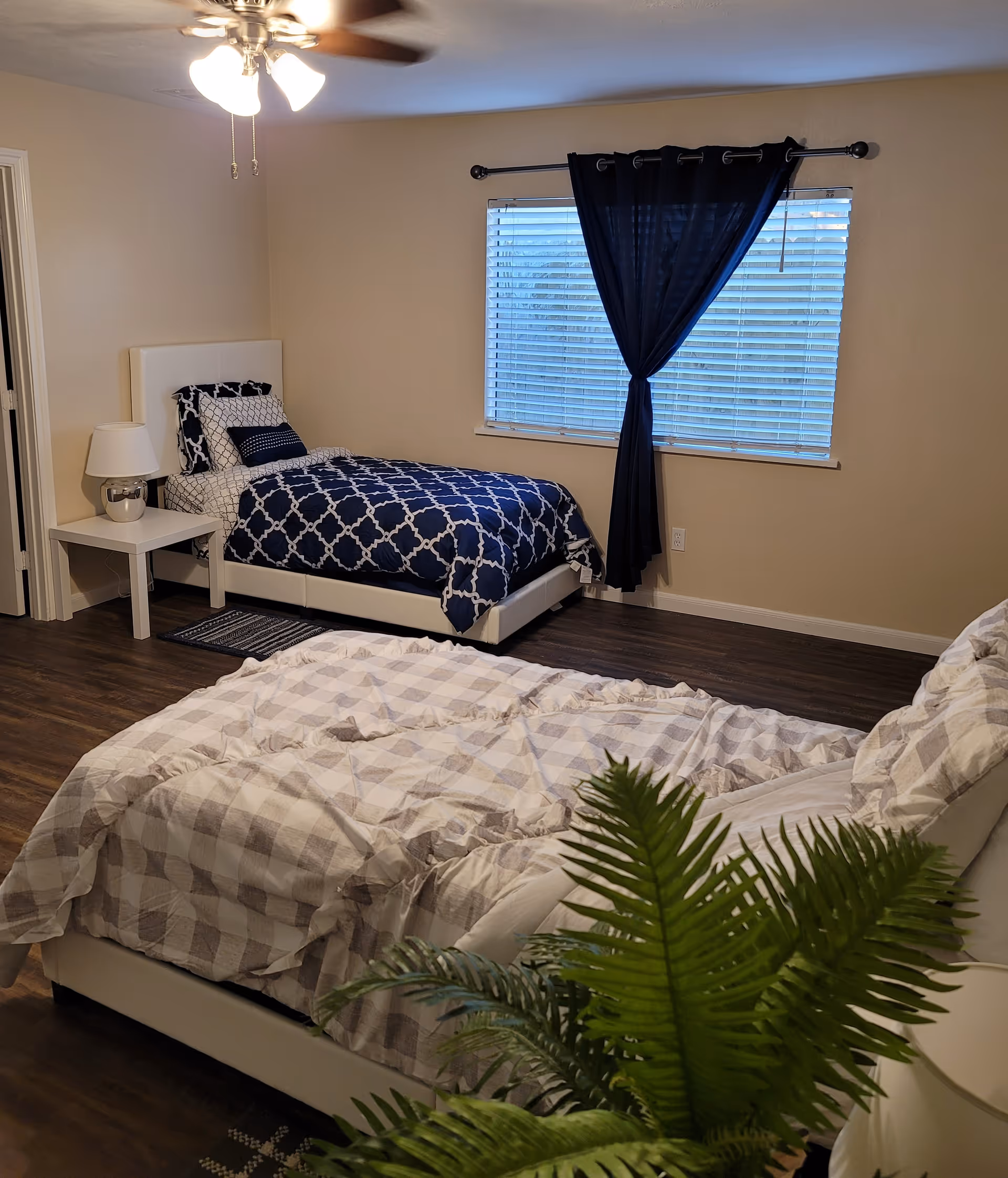 A bedroom with two beds, one with a blue and white patterned comforter and the other with a beige and white checkered comforter. There is a white nightstand with a lamp next to the bed with the blue comforter. A window with closed blinds and a dark curtain is on the wall behind the beds. A ceiling fan with lights is on the ceiling, and a green plant is partially visible in the foreground.