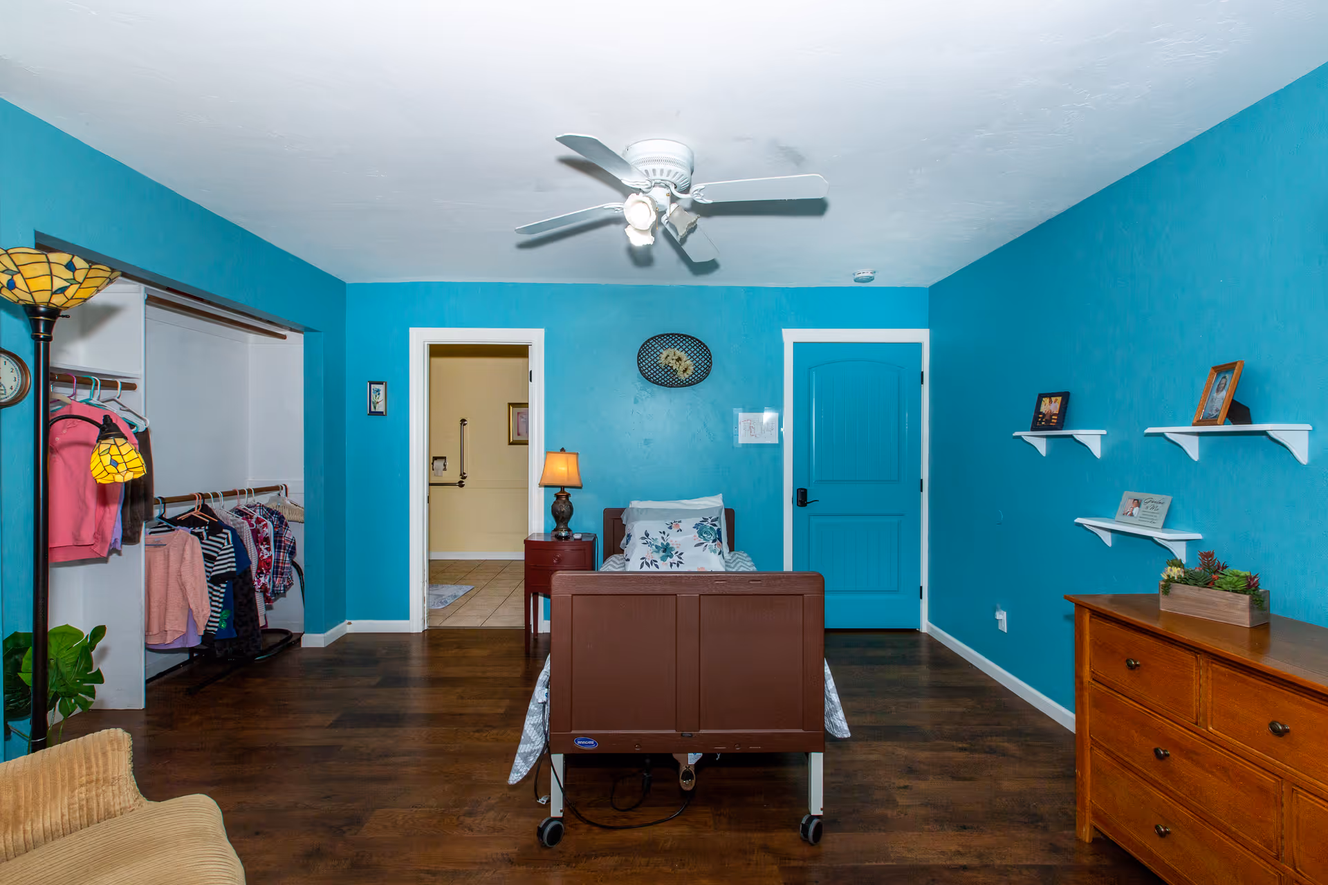 A bedroom in Prescott Valley Assisted Living with teal blue walls and dark wood flooring. The room features a hospital-style bed with floral bedding, a wooden dresser, a nightstand with a lamp, and an open closet with hanging clothes. There are three small white shelves on the right wall with framed photos and a small plant. A ceiling fan with lights is mounted on the white ceiling. The room has two doors, one leading to a bathroom and the other closed and painted teal blue.