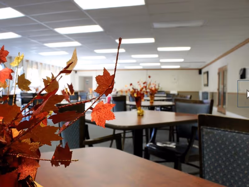 Interior view of a dining room with multiple tables and chairs arranged neatly. Each table has a centerpiece with autumn-themed decorations featuring red and orange leaves. The room is well-lit with ceiling lights and has a calm, welcoming atmosphere.