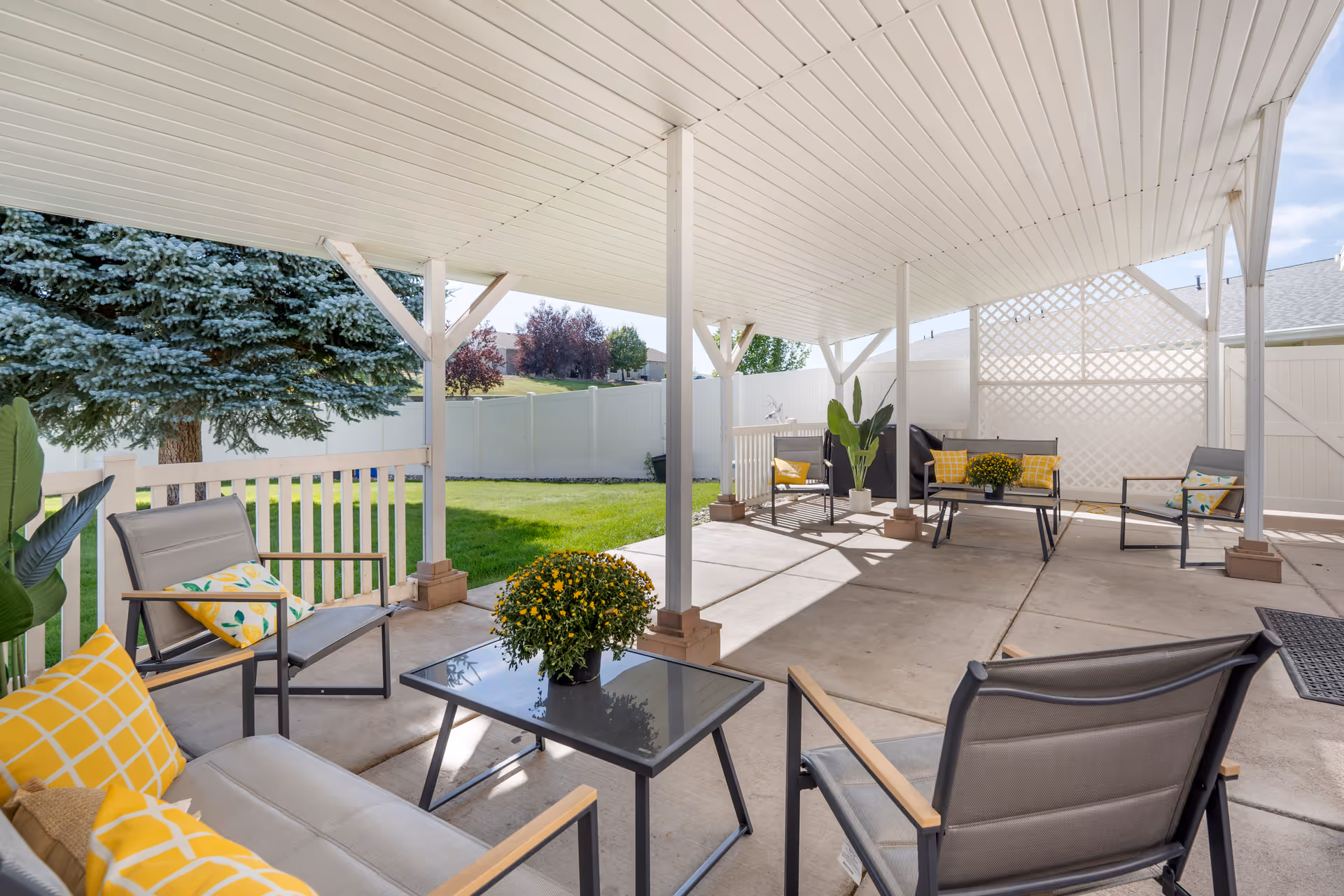 Covered outdoor patio area with several metal-framed chairs and a sofa with yellow and patterned cushions, a glass coffee table with a potted plant, and a white fence and green lawn in the background.