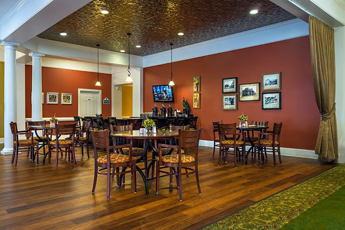 A dining area with several wooden tables and chairs with patterned cushions. The room has a warm, reddish-brown wall adorned with framed pictures and a mounted TV. There are pendant lights hanging from a decorative ceiling, and a counter with coffee dispensers in the background. The floor is wooden with a green carpeted edge on one side.