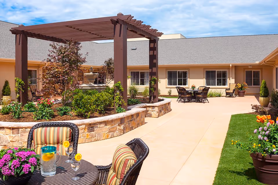 Outdoor courtyard area at Lakeview Memory Care featuring a pergola over a stone planter with greenery and a fountain, multiple seating areas with wicker chairs and tables, colorful flowers in pots, and a clear blue sky.