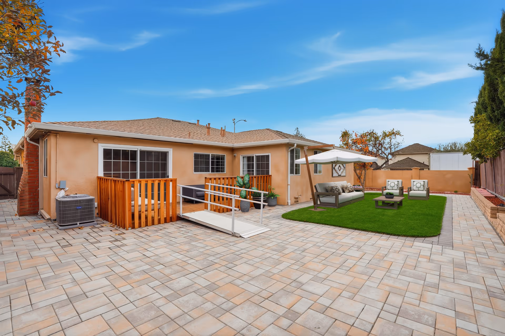 Sunny backyard with tiled patio, wooden deck, and outdoor seating on artificial grass.