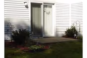 Exterior view shows a white siding building entrance with a glass door and shrubs.