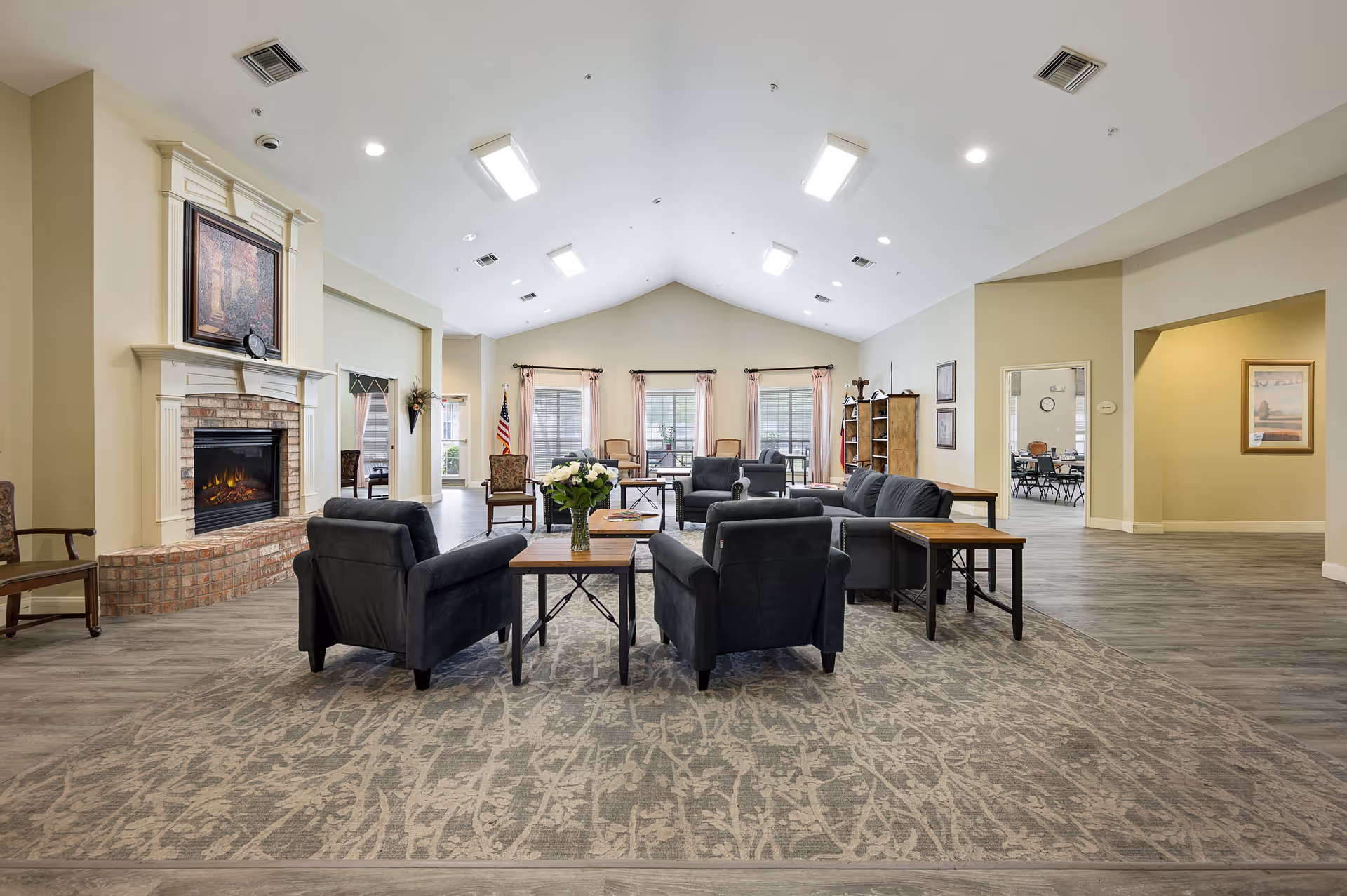 Spacious senior living facility common area with high vaulted ceilings, multiple black armchairs and sofas arranged around wooden coffee tables on a patterned carpet. A brick fireplace with a white mantel and framed artwork above is on the left wall. Large windows with sheer curtains allow natural light into the room. An American flag is visible near the windows, and a doorway leads to another room with tables and chairs.