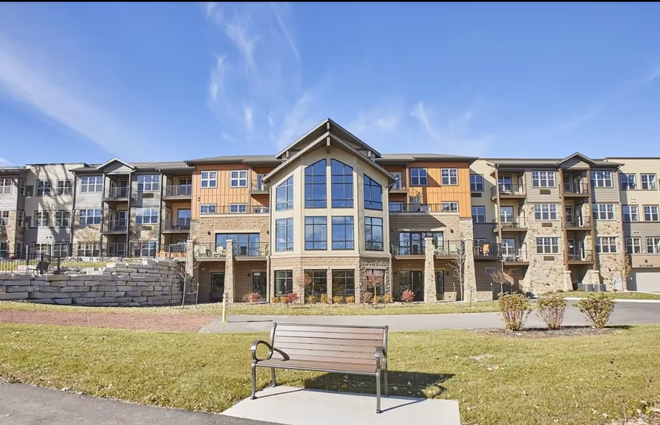Front exterior of a multi-story senior living building with a large central glass facade, grassy lawn, and a bench in the foreground.