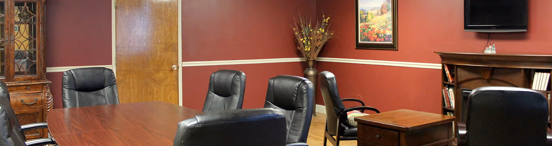 A conference room with a large wooden table, black leather chairs, and maroon walls.